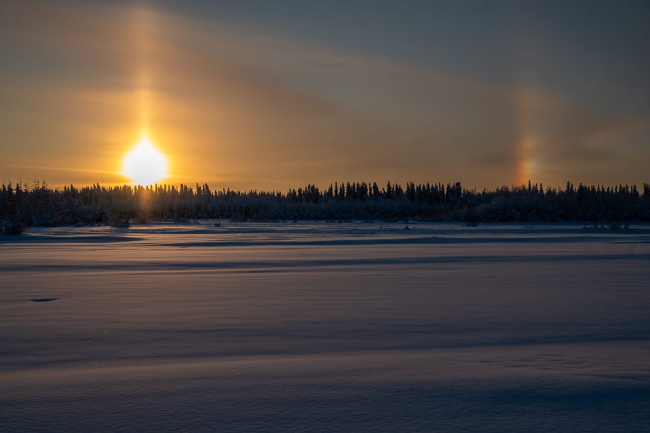 A bright winter sun glows low above a distant forest beyond a wide, frozen lake. A strong vertical sun pillar rises above the sun, and a colorful sundog appears to the right, while the snow-covered lake in the foreground reflects faint warm light. A bright winter sun glows low above a distant forest beyond a wide, frozen lake. A strong vertical sun pillar rises above the sun, and a colorful sundog appears to the right, while the snow-covered lake in the foreground reflects faint warm light.