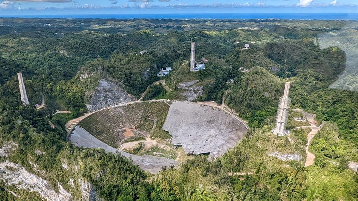 LEFT: Arecibo Observatory is set in forested mountains with the concrete bowl contrasting verdant green. Wires are run between three tall supports strung like a suspension bridge with an array of equipment dangling between the three on lines. RIGHT: The Arecibo Telescope during demolition process, December 2021. Credit: Tedder at Wikimedia Commons (https://commons.wikimedia.org/wiki/File:Arecibo_Telescope,_December_2021.jpg)