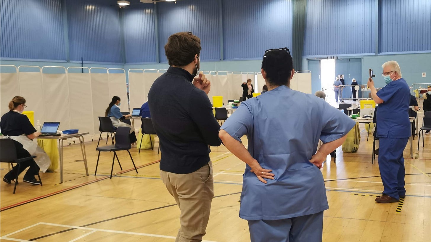 View of some a leisure centre sports hall turned into a temporary vaccination centre. There are people sitting at workstations with open laptops in front of them. 