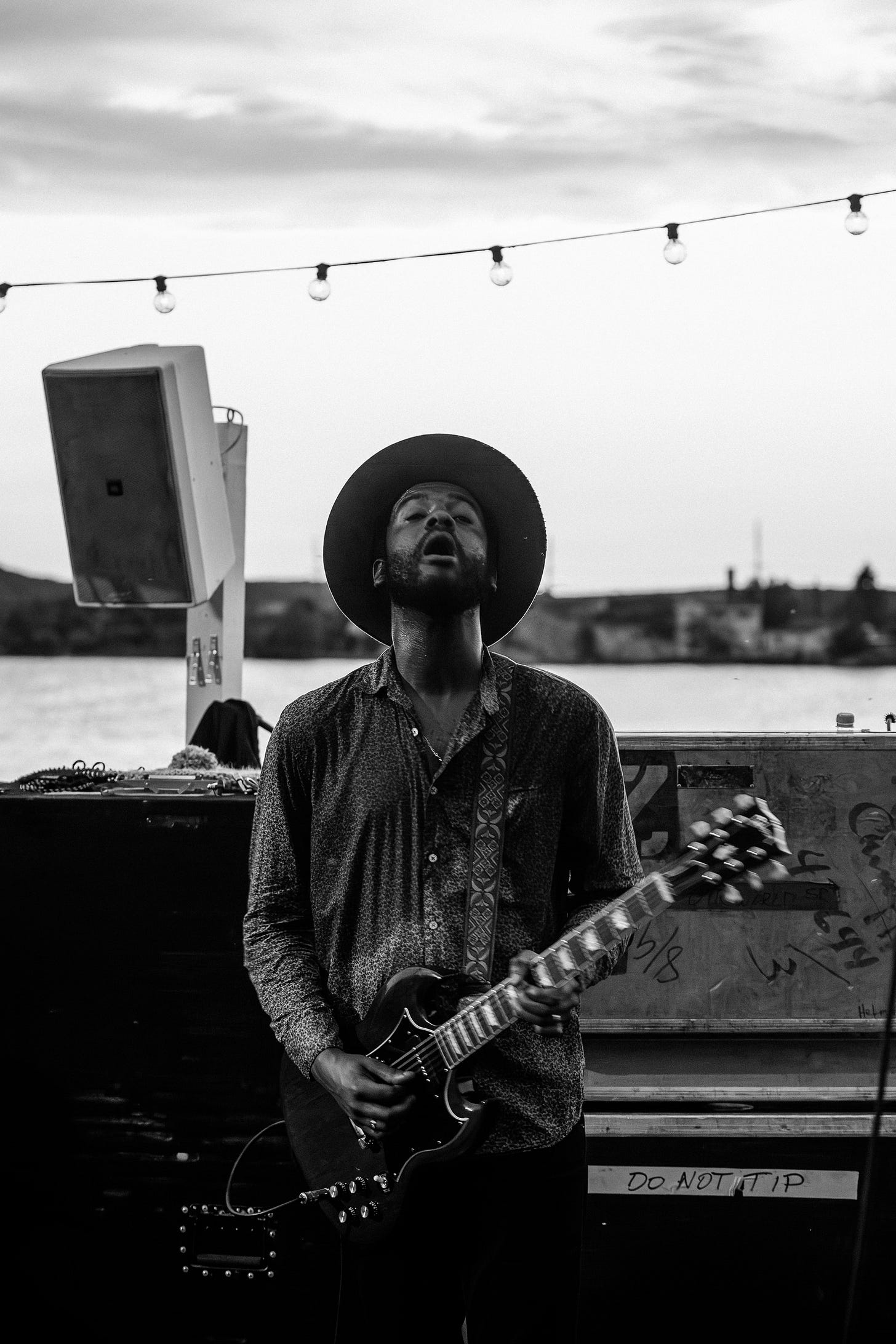 A black and white photo of Gary Clark Jr. looking up with emotion while playing his electric guitar on an outdoor stage. String lights are visible above him, and water is in the background.