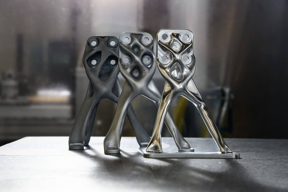 Three similar parts sit on a table. Organic-looking scaffolding crosses over the front