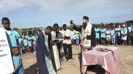 Un évêque orthodoxe, avec barbe et bure noire, donne l'onction à un jeune enfant malgache, sous le soleil écrasant de Madagascar