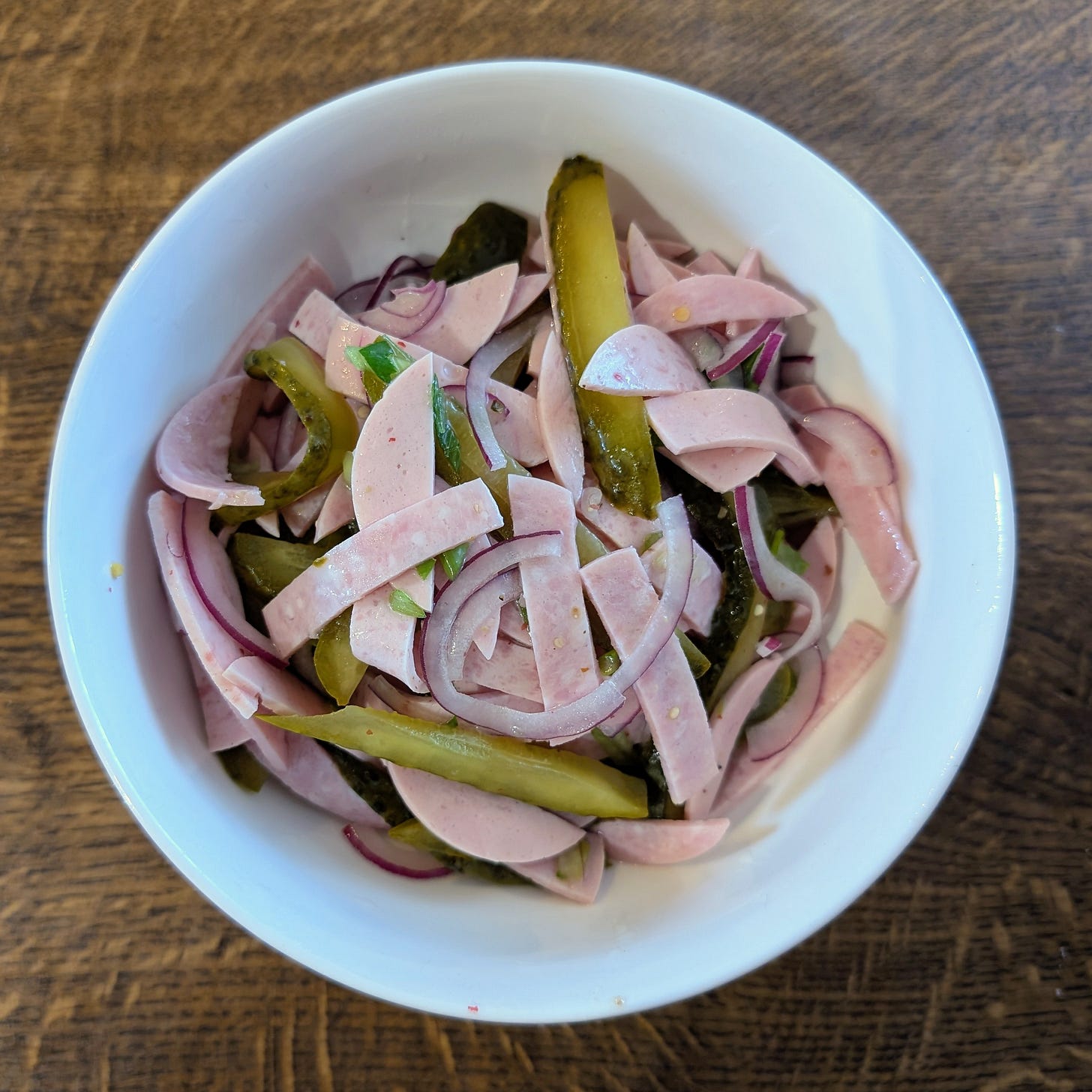 A white bowl filled with a German-style Wurstsalat made of thinly sliced sausage, red onion rings, and sliced pickles, all mixed together. The bowl sits on a wooden surface, showing the salad’s mix of pink, green, and purple tones.