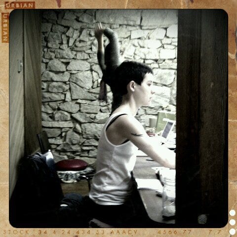 Author seated at a desk in Chinese Medicine school classroom, wearing a white tank top with short shaved hair, focused on studying.
