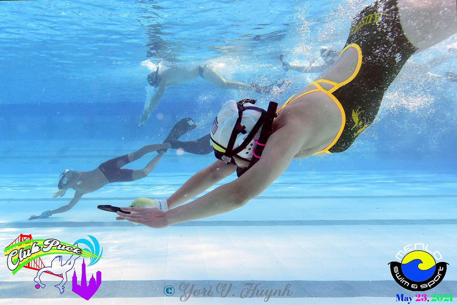 3 people diving towards pool floor; each wearing fins, a swimsuit, snorkel with polo cap, and holding a stick in a gloved hand.