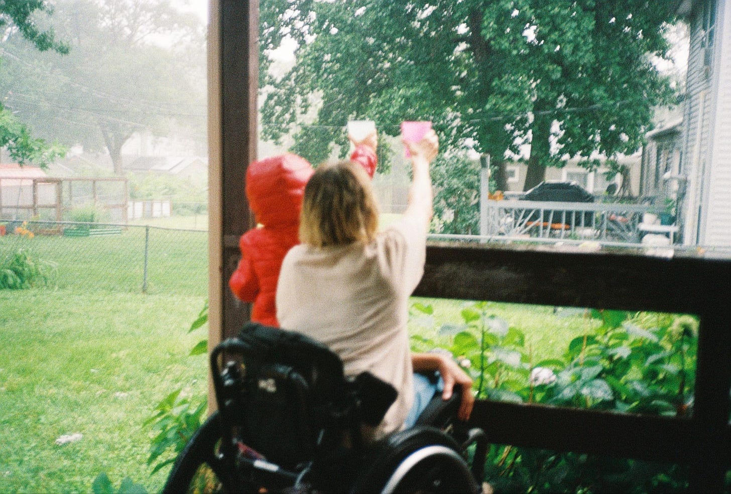 Rebekah and her son hold their cups up to the rainy sky again. This time they're under the shelter of a deck, and the photo is taken from behind. The yard around them is green and misty.