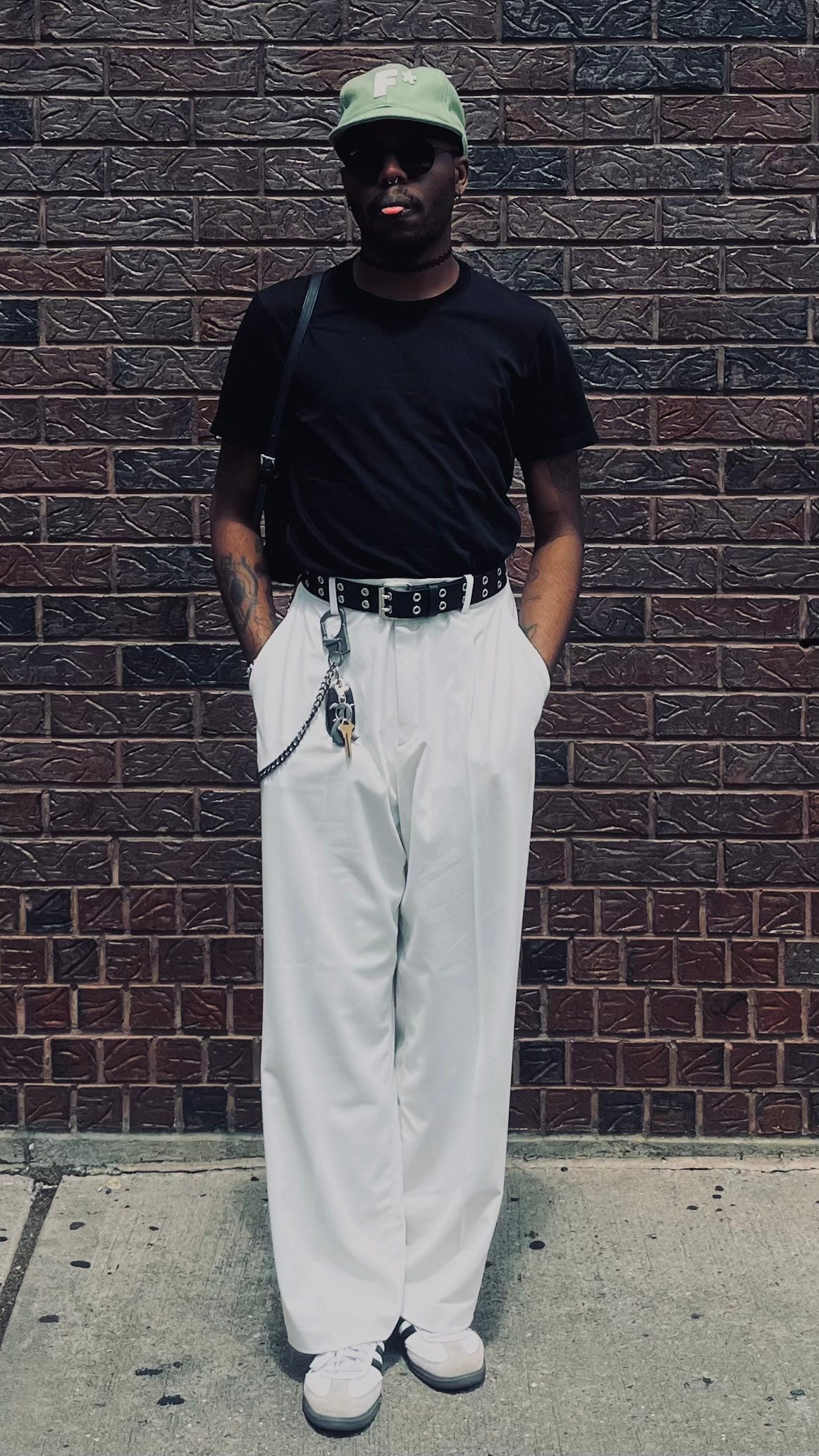 elle slow stands in front of a brick wall, tongue slightly out, with a pastel green golf le fleur hat, black tee shirt, vintage burberry purse, off-white pleated wide-leg trousers, and white-and-black adidas sambas.