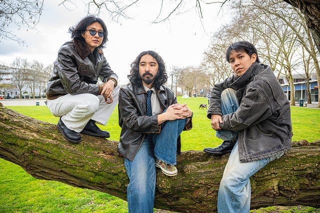 Three Asian men sitting on a large tree branch with yellow-green grass behind them