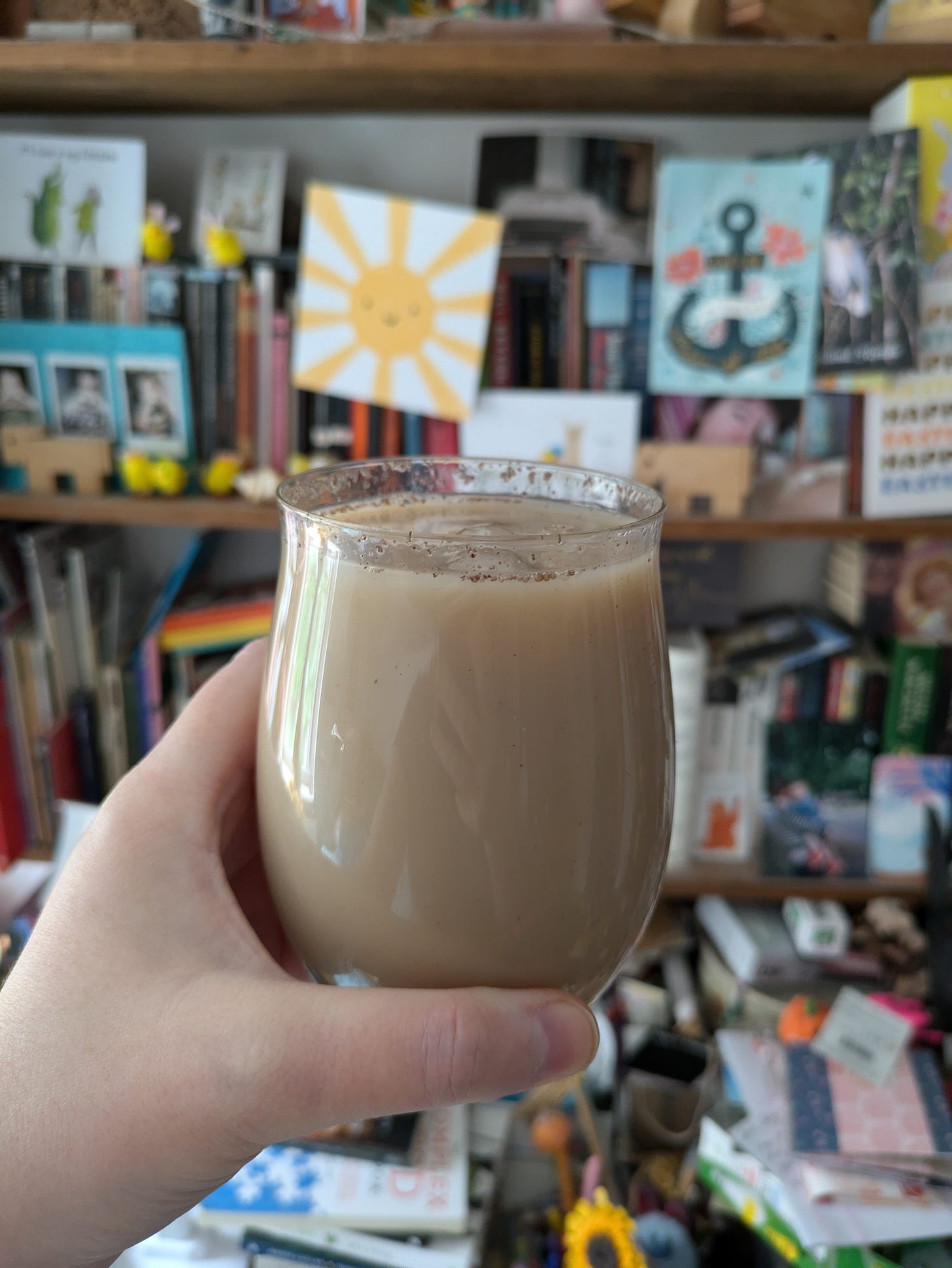 ID. A photo of my hand holding a nicely shaped glass full of iced chai in front of a blurred background of a colourful bookshelf with lots of cards/photos/trinkets. ID. A photo of my hand holding a nicely shaped glass full of iced chai in front of a blurred background of a colourful bookshelf with lots of cards/photos/trinkets.