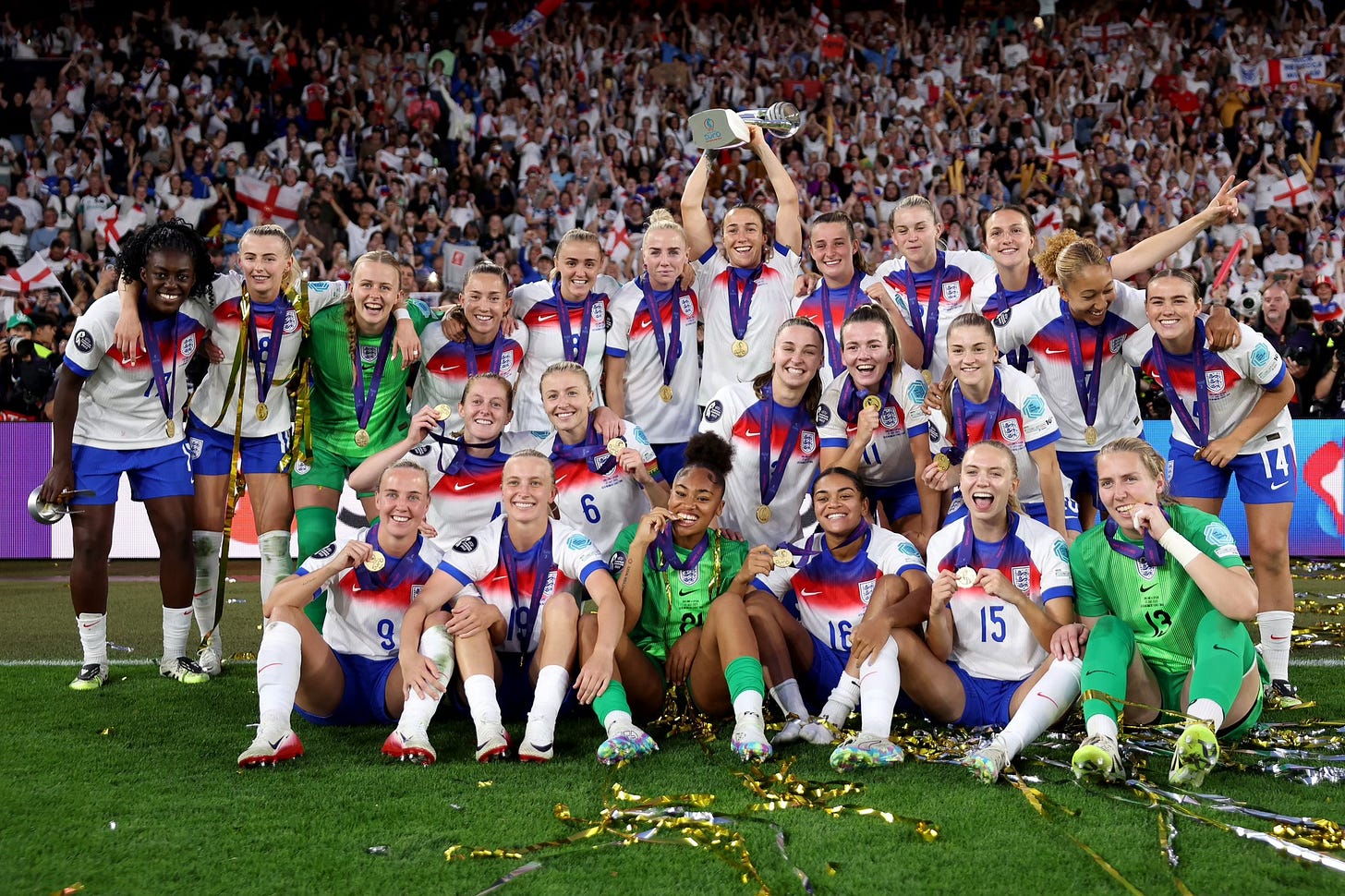 The England women’s national football team poses together on the pitch, celebrating their victory with medals and a trophy after winning a major tournament. The players wear white and blue kits with red accents and are smiling and cheering, surrounded by gold confetti. One player stands in the centre holding the trophy aloft, while others kneel or crouch in front, holding up their medals. A large crowd of fans with England flags fills the stadium in the background.