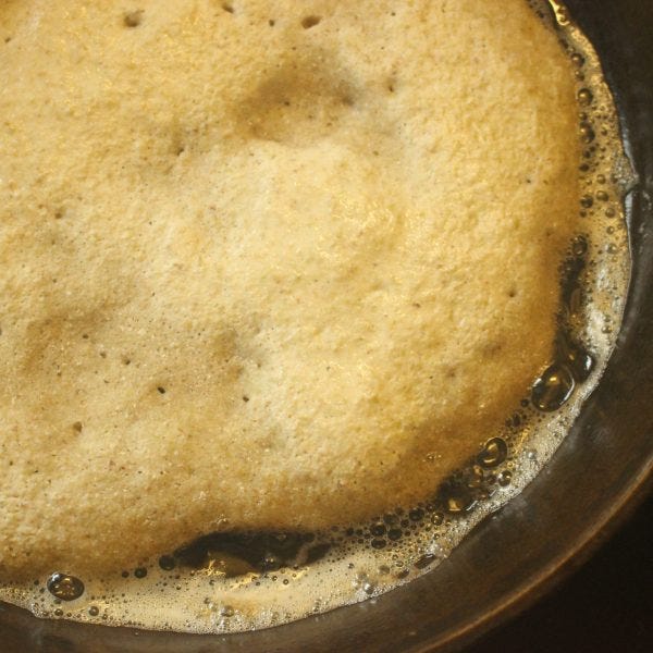 shows about a quarter of a dark pan filled with a fying ancient grain pancake with bubbles before it has been flipped. Bubbling oil at the edges. 