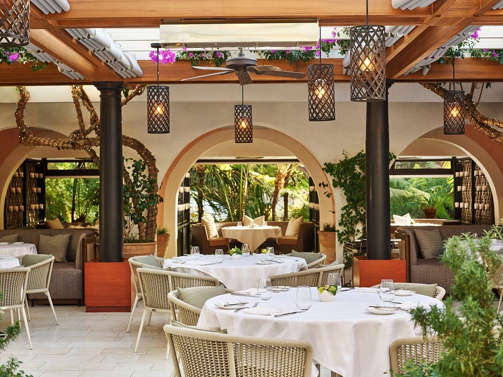 Luxurious restaurant patio with round tables draped in white cloth, wicker chairs, and warm, woven lanterns. Arched windows reveal lush greenery outside.