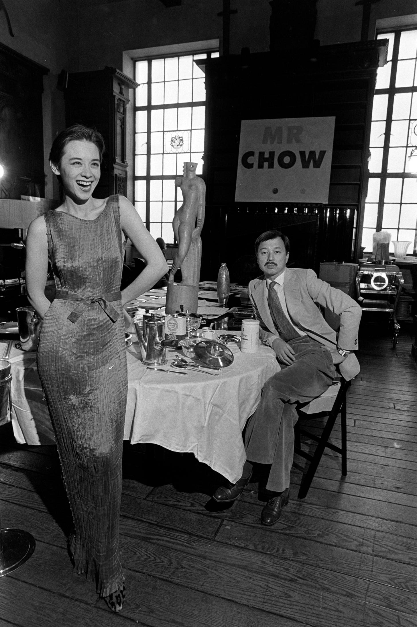 A woman wearing a sleeveless silk dress stands in front of a restaurant dining table while a suited man sits at the table with a somber expression.