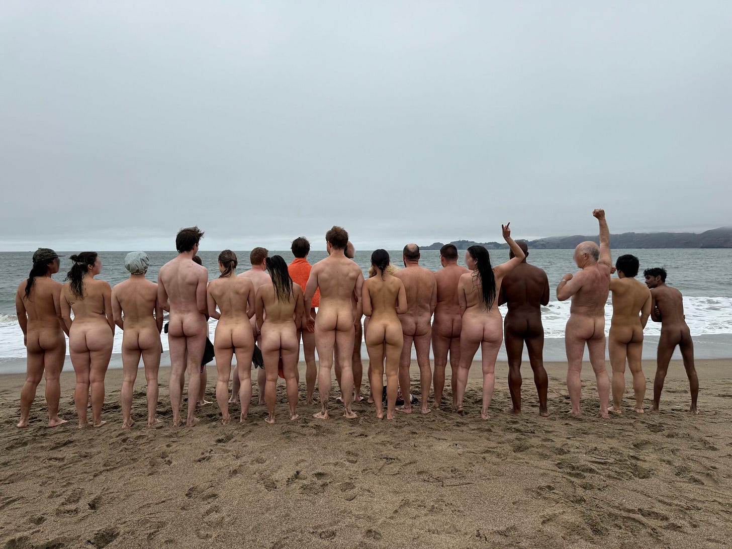 A group of nude people stand in a line facing the ocean on a cloudy day at Baker Beach in San Francisco. Some raise their arms in celebration. A group of nude people stand in a line facing the ocean on a cloudy day at Baker Beach in San Francisco. Some raise their arms in celebration.
