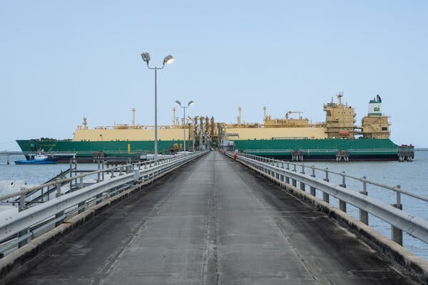 A green-and-yellow L.N.G. shipping tanker is parked in the water. There is a long concrete pier leading to the tanker. A green-and-yellow L.N.G. shipping tanker is parked in the water. There is a long concrete pier leading to the tanker.