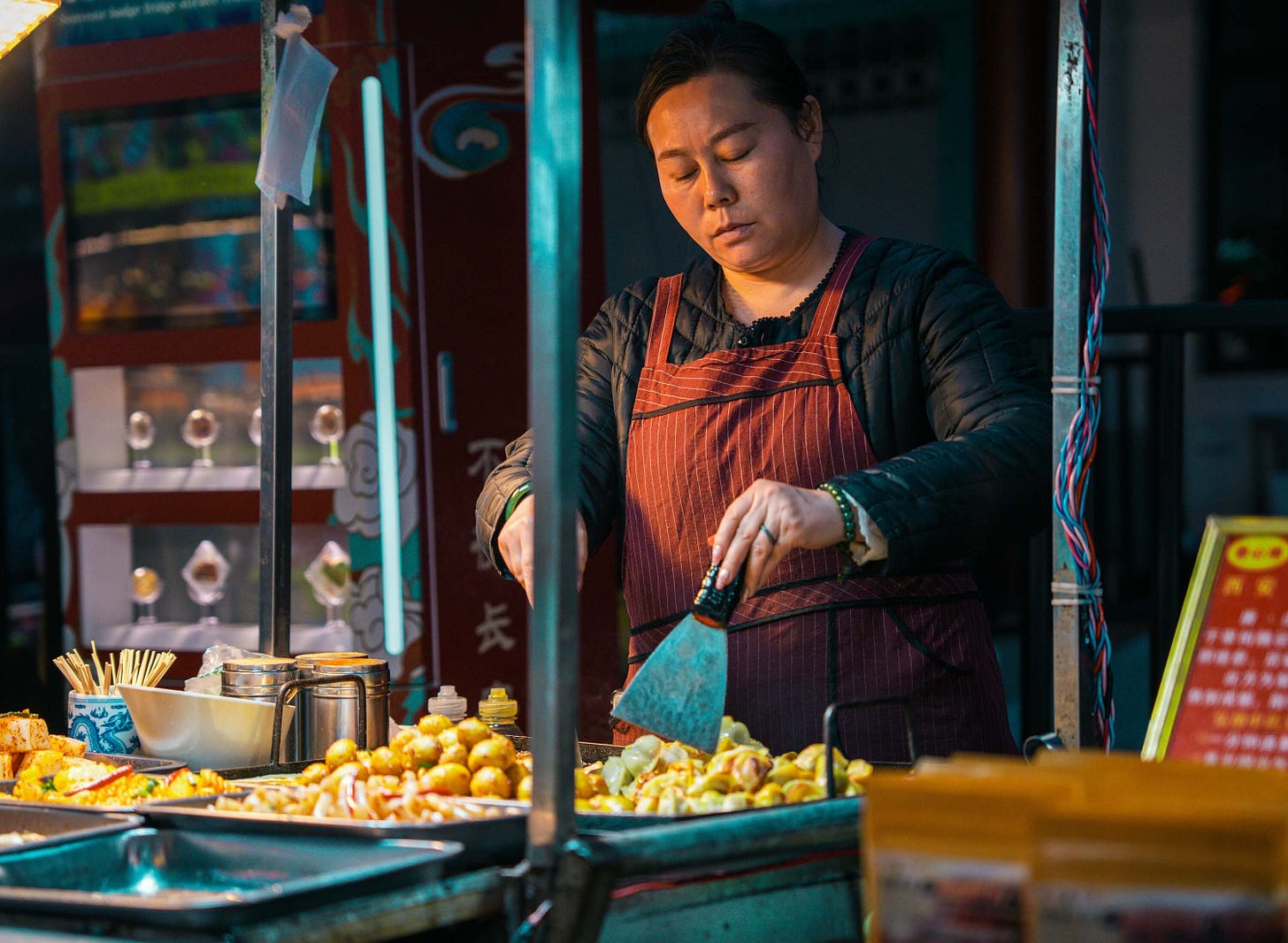 A woman cooks dumplings at her market stall in Xi’an in Shaanxi province, central China. She wears a black shirt and red apron and there's a blue light from her metal stand. © Sergio Kian 2025 A woman cooks dumplings at her market stall in Xi’an in Shaanxi province, central China. She wears a black shirt and red apron and there's a blue light from her metal stand. © Sergio Kian 2025