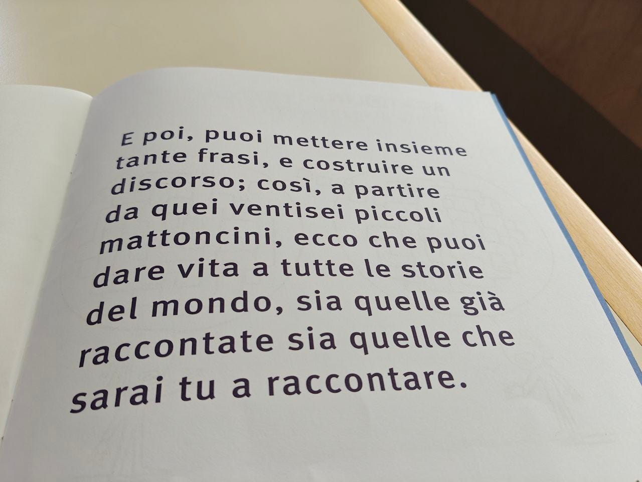 Una pagina della parola PAROLA delle Scatoline di effequ, con questa scritta: "E poi, puoi mettere insieme tante frasi, e costruire un discorso; così, a partire da quei ventisei piccoli mattoncini, ecco che puoi dare vita a tutte le storie del mondo, sia quelle già raccontate sia quelle che sarai tu a raccontare". La pagina è appoggiata su un tavolo di legno bianco sporco e nell'angolino in alto a sinistra si intravede un pezzetto di muro in ombra.