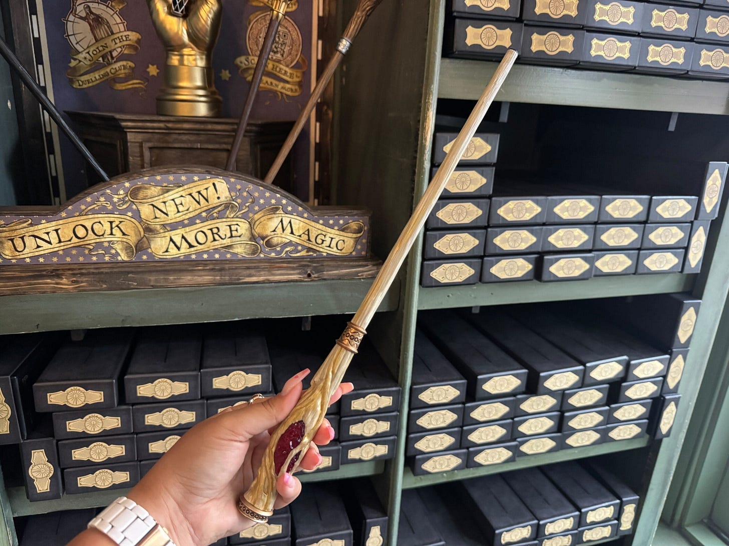 A hand holds a decorative wand in a shop filled with wand boxes at Universal Orlando Resort. A sign reads, "Unlock More Magic." With second-gen interactive wands, the experience is simply enchanting.