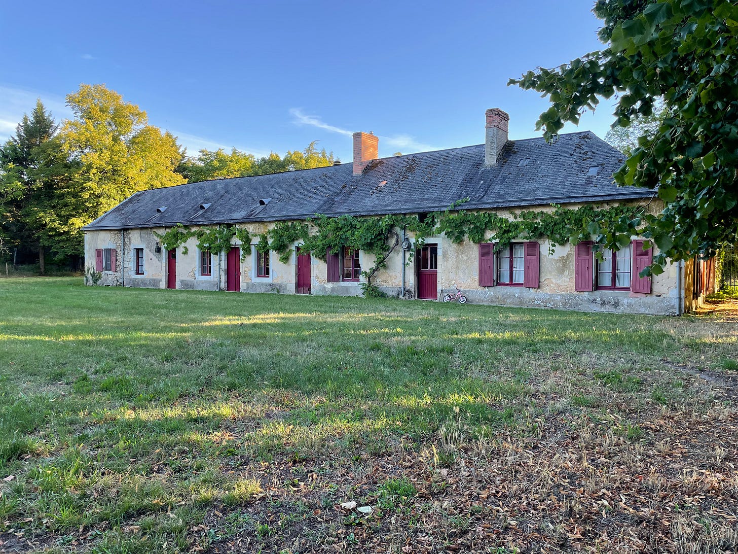 A long, low stone farmhouse with a slate roof, climbing vines, and deep red shutters, sitting at the edge of a wide lawn with a child's pink bicycle parked by one of the doors. A long, low stone farmhouse with a slate roof, climbing vines, and deep red shutters, sitting at the edge of a wide lawn with a child's pink bicycle parked by one of the doors.