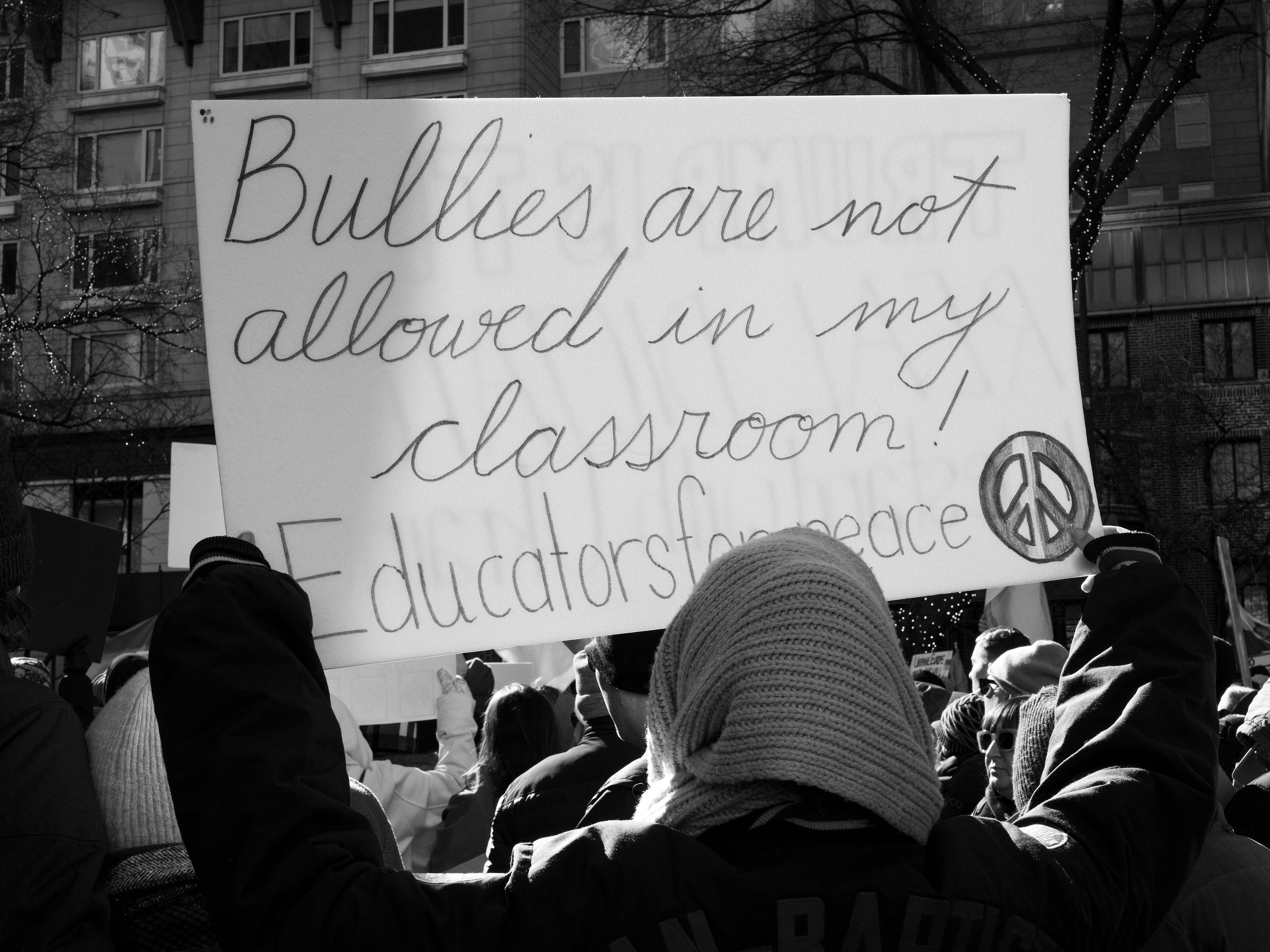 Bullies are not allowed in my classroom! #Educatorsforpeace," accompanied by a peace symbol. The person, wearing a knitted beanie and winter coat, stands among a crowd of demonstrators. The background features urban buildings, trees adorned with string lights, and other protesters holding signs, emphasizing the collective stand against injustice. Bullies are not allowed in my classroom! #Educatorsforpeace," accompanied by a peace symbol. The person, wearing a knitted beanie and winter coat, stands among a crowd of demonstrators. The background features urban buildings, trees adorned with string lights, and other protesters holding signs, emphasizing the collective stand against injustice.