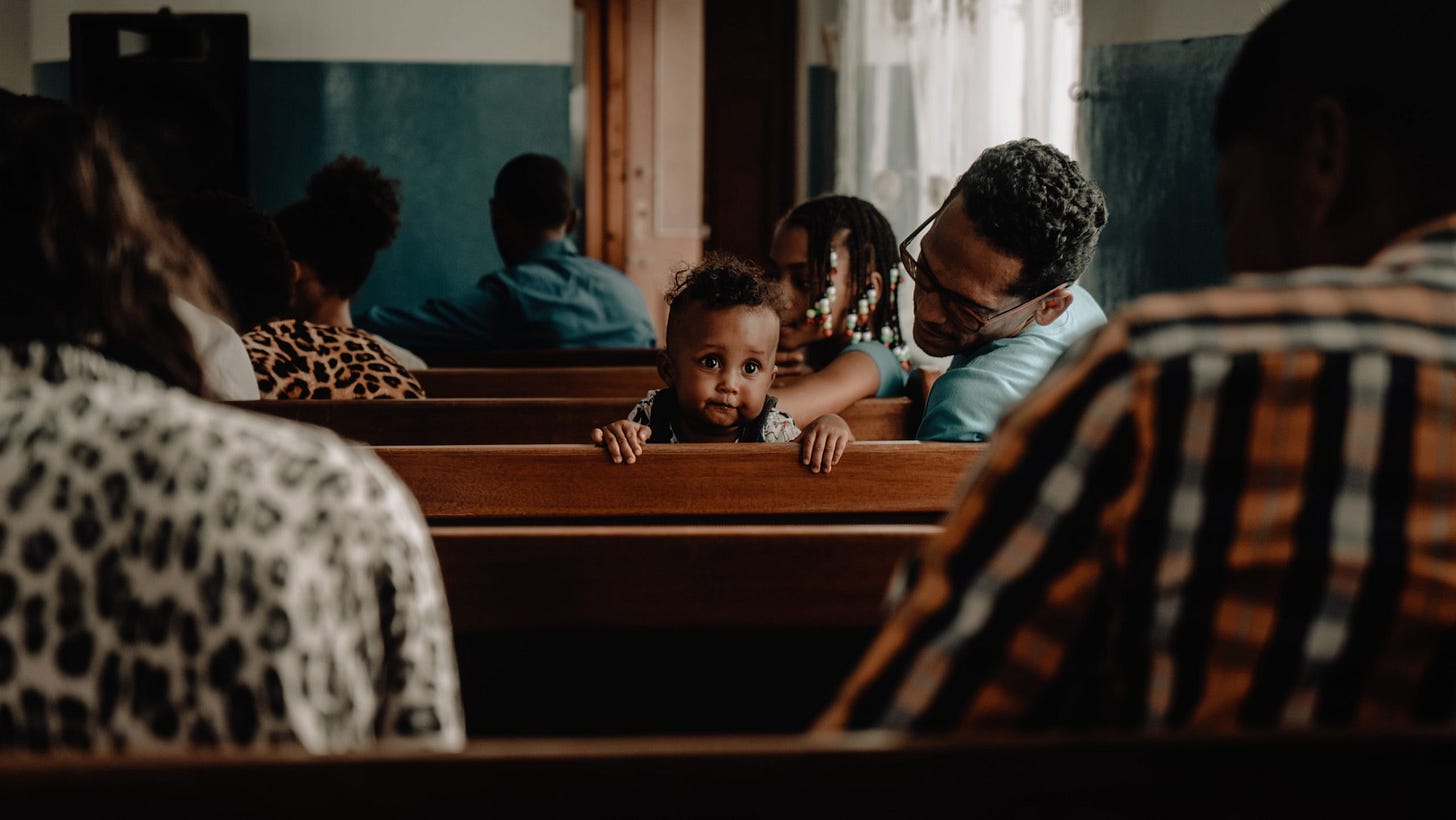 People sit in views with the backs of their head visible. In one pew at the center of the image, a baby turns around and looks back at the viewer.
