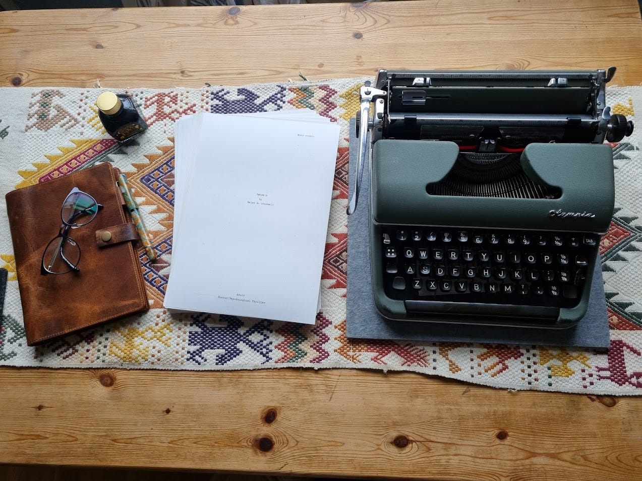 A typewriter, a leather notebook and a manuscript of a book on a woven table runner on a wooden table. The book is called "Twine'd by Helen R Gradwell."