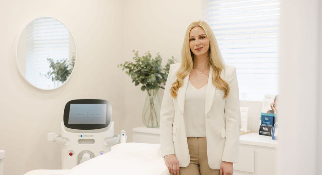 Dr Jess - a blonde woman wearing a white blazer and beige trousers - standing in front of a clinic bed and sofwave machine
