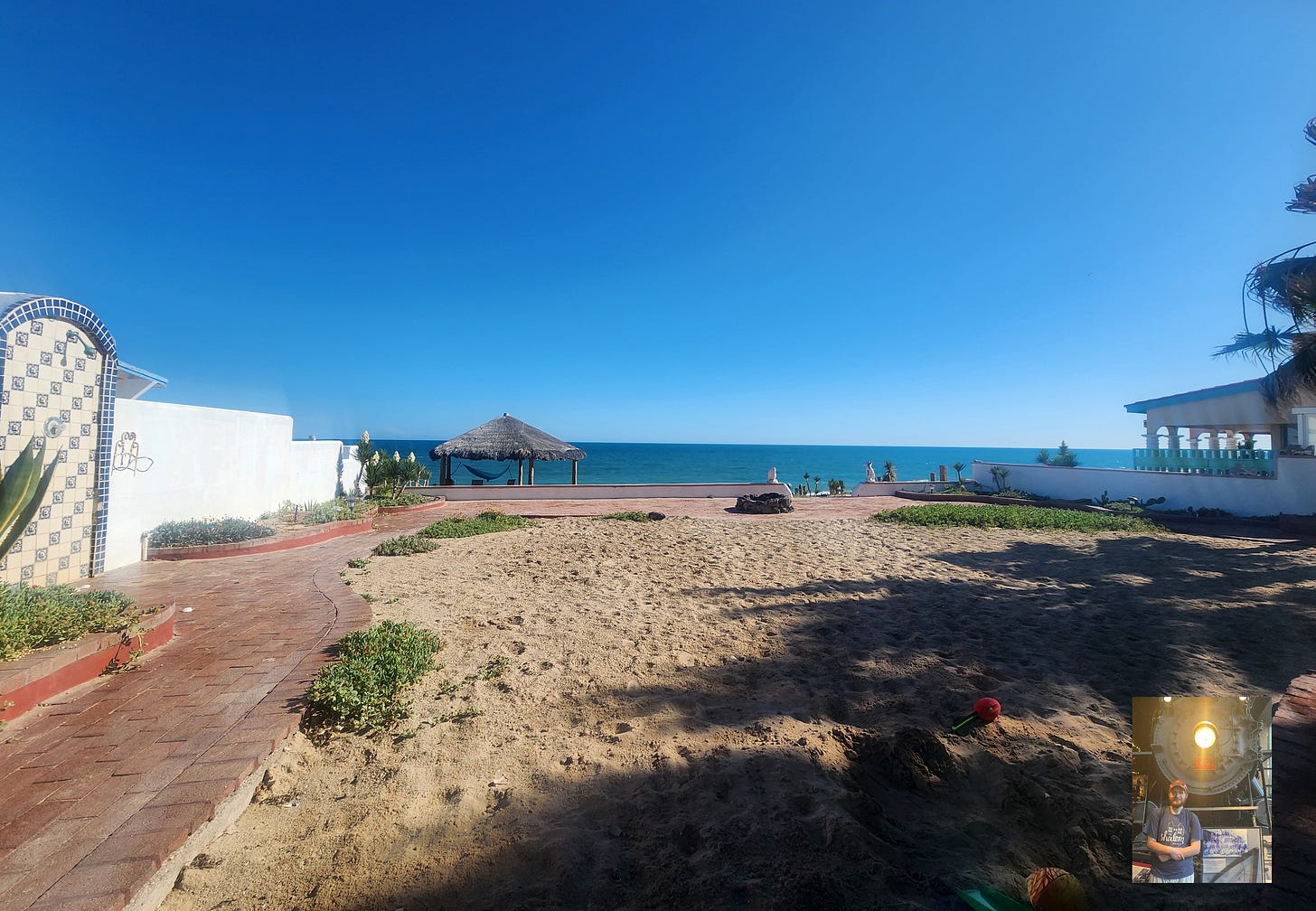 An ocean facing view of a house in Agua Prieta, Mexico. To the left along a white wall is an outdoor shower, and follows a brick path. In the background is a sand yard. In the background is the Pacific Ocean.