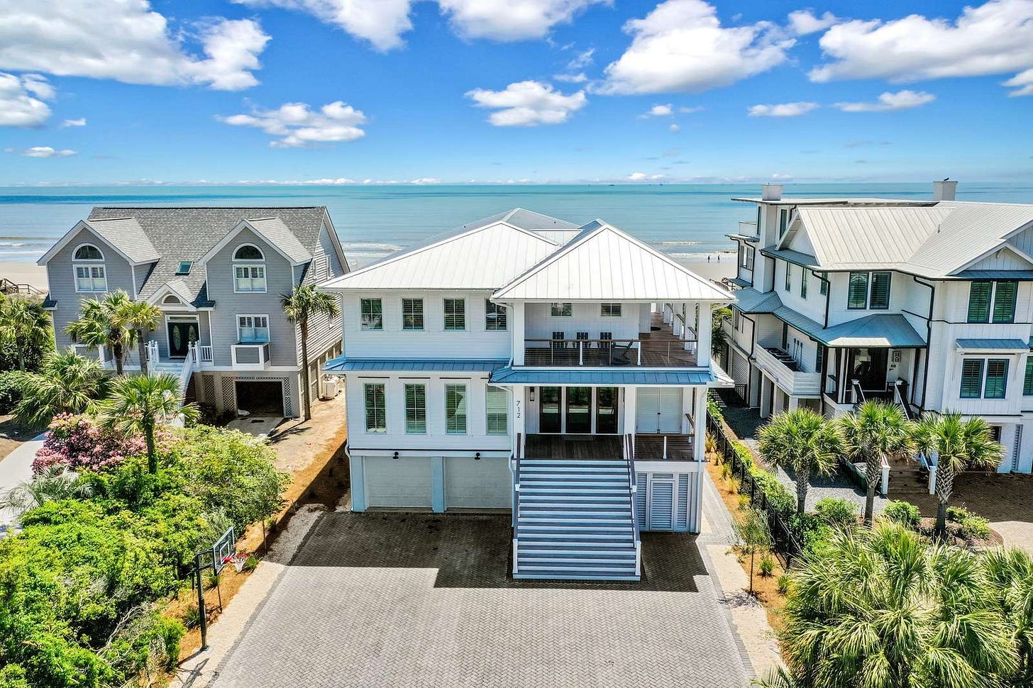 Aerial view of the front of a white beach house with a metal roof and a wide staircase up to the entry, with the ocean visible directly behind it. Aerial view of the front of a white beach house with a metal roof and a wide staircase up to the entry, with the ocean visible directly behind it.