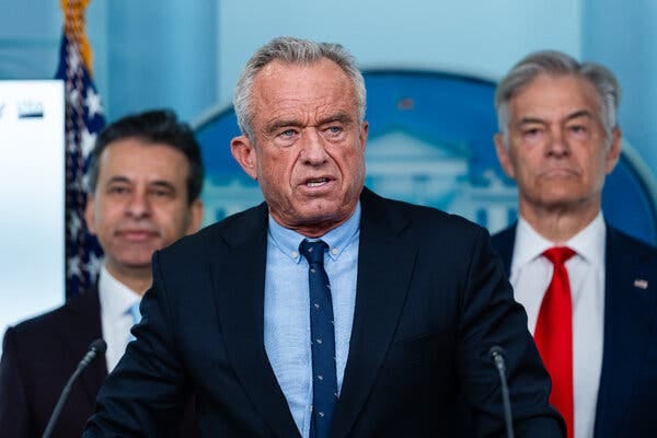 Health Secretary Robert F. Kennedy Jr., in a dark suit and a blue patterned tie, stands between two other men.