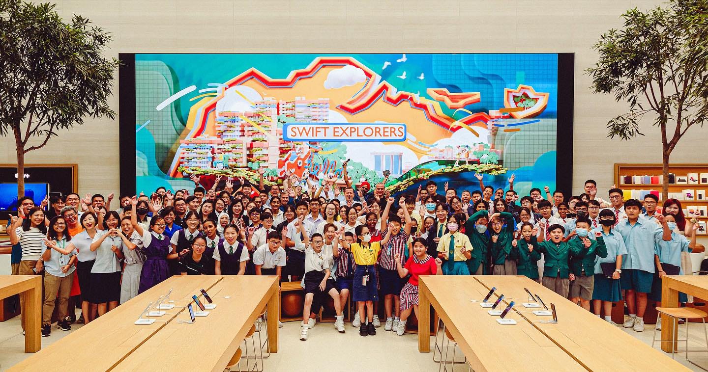 A large group of students stand in front of the video wall at Apple Orchard Road. A Swift Explorers animation is playing on the wall. A large group of students stand in front of the video wall at Apple Orchard Road. A Swift Explorers animation is playing on the wall.