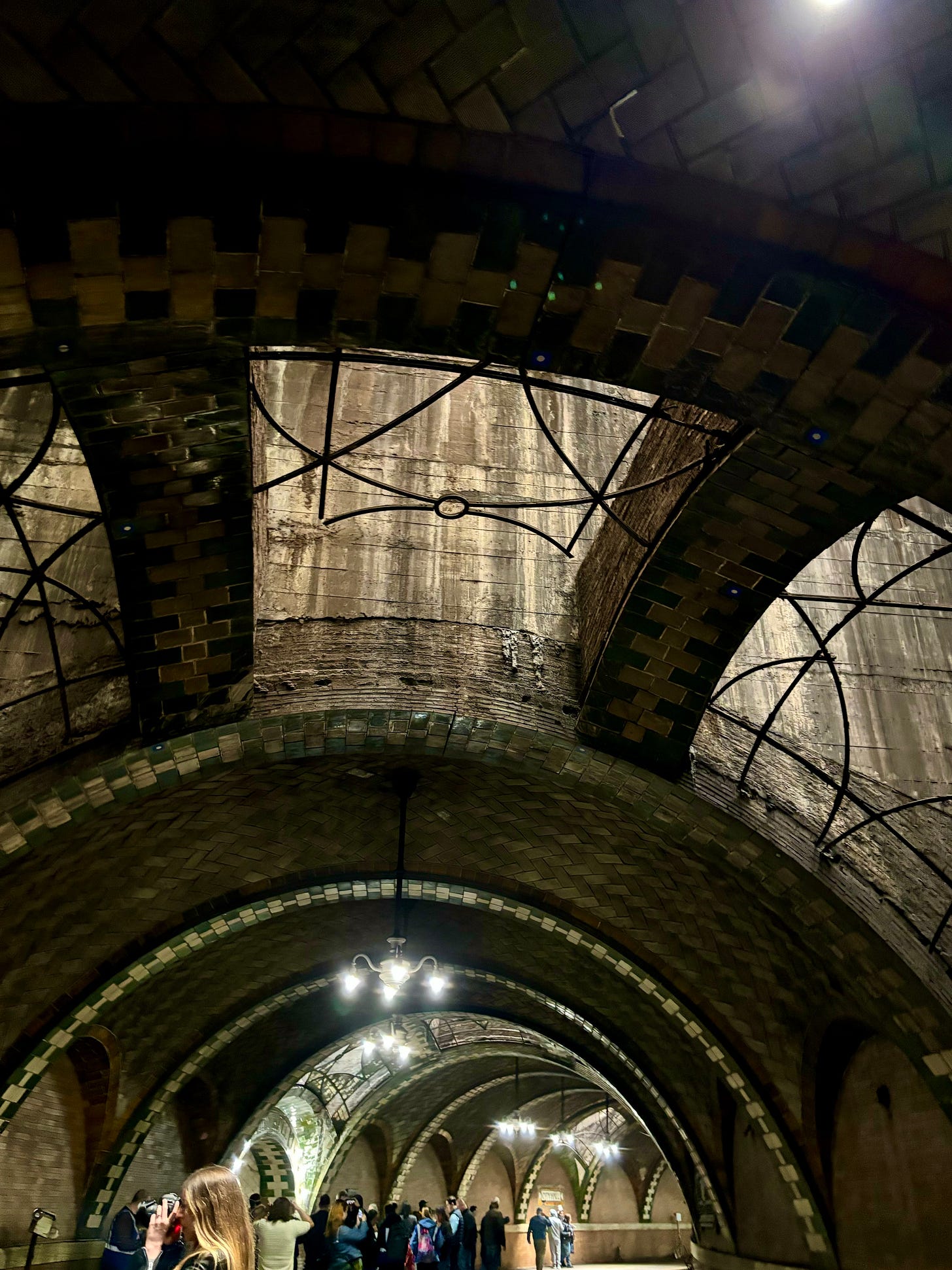 Sunlight coming in through the windows in the arched ceiling looking down the station where the tiled arches and chandeliers are visible. People can be seen taking pictures of the station.