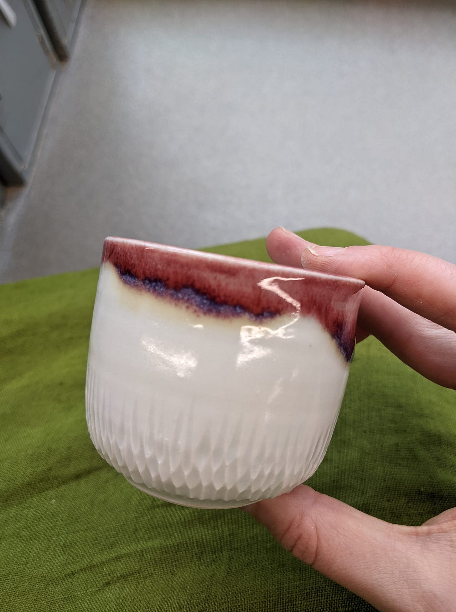 A celadon cup with red flashed on top, and little bits of clay removed from the bottom. The background is a moss green linen apron.