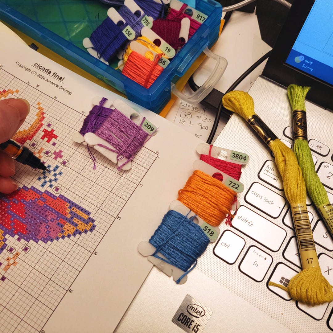 A disheveled workspace showing a laptop keyboard, a printed cross stitch pattern, and loose embroidery skein and floss bobbins.