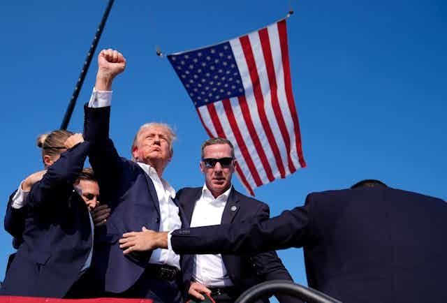 A man with greyish blond hair and a streak of blood on his face raises his fist in the middle of a huddle of Secret Service agents. An American flag flies next to him.