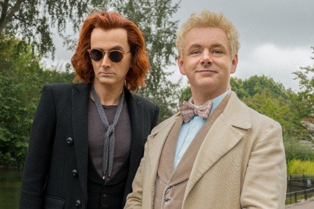 A picture of two male-shaped beings standing side by side. On the left is a lanky man with shoulder-length red hair, round sunglasses, and all black attire. On the R is a shorter man in a tan and cream three piece suit. He has short, curly blond hair.