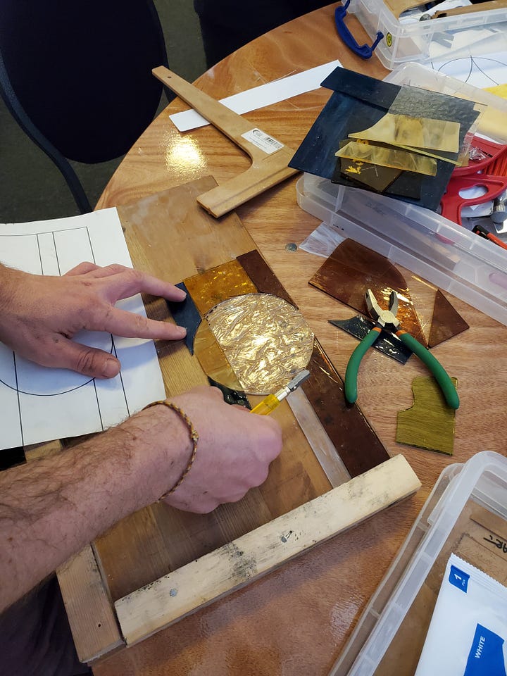 First Image: A person is working on a stained glass project. Their hands are carefully arranging pieces of colored glass, including yellow, gold, and black sections. Tools like pliers and a cutting tool are visible on the table, alongside various sheets of glass and a wooden frame. There is a paper design outline partially shown underneath the glass pieces. Second Image: A partially completed stained glass artwork is in progress, showing a colorful sunburst design. Green and yellow glass rays emanate from an orange half-sun, with blue and green glass pieces arranged at the bottom. The design is laid out in a wooden frame, with scattered glass pieces and tools like clamps, pliers, and a cutting tool visible around the workspace.