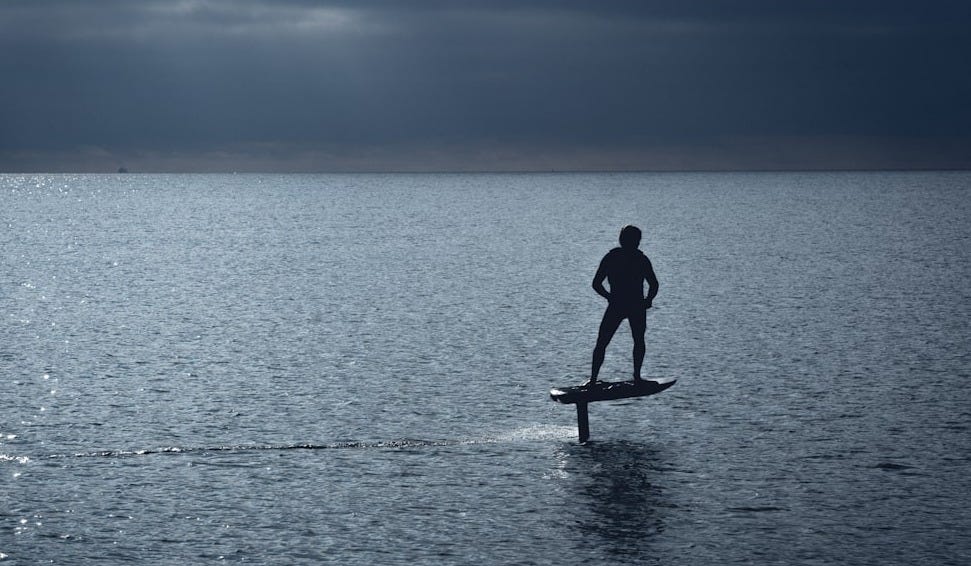 man in black wet suit standing on brown wooden paddle board during daytime man in black wet suit standing on brown wooden paddle board during daytime