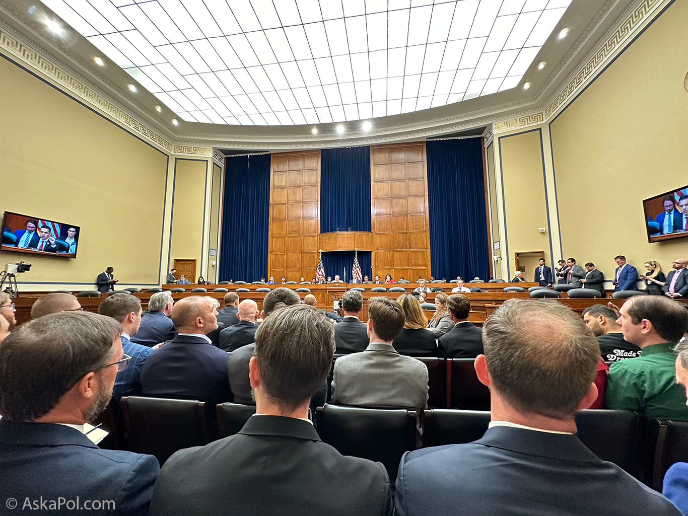 Men in suits sit listening in expansive room as people on dais speak. Photo: Matt Laslo © www.askapol.com