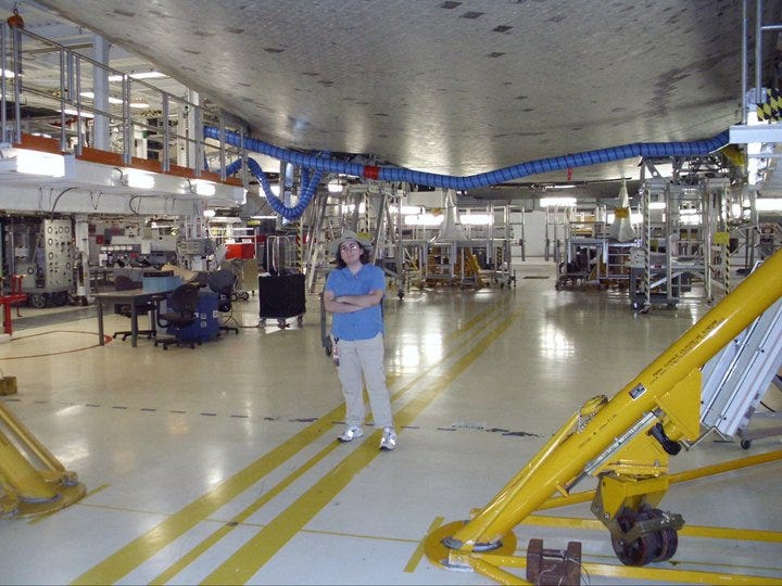 The author stands underneath the shuttle Endeavour while it's docked at the OPF. The shuttle is held up by scaffolding and big lifts (not unlike one would use to change a tire). The belly of the shuttle is tiled, and some tiles are darker than others. It looks like the roof of a short building.