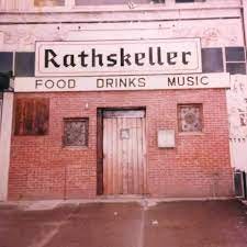This is a photograph of the front entrance of a building with a red brick facade. A large sign spanning the top of the building reads "RATHSKELLER" in a Gothic font. Below this, a smaller sign indicates "FOOD DRINKS MUSIC." The entrance features a large, rustic wooden door in the center, flanked by two small, barred windows. The overall appearance is worn and suggests an older, no-frills establishment.