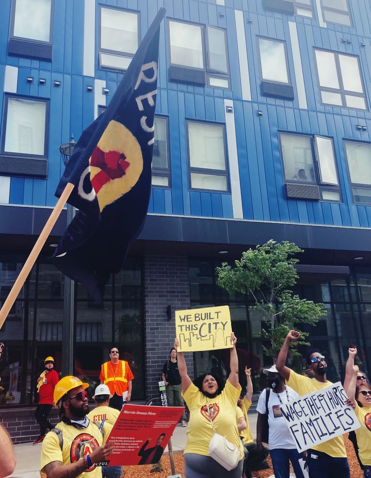 people in bright yellow shirts hold up signs, one reading "we built this city" and another person carrying a dark flag, some letters of the word "resist" are visible