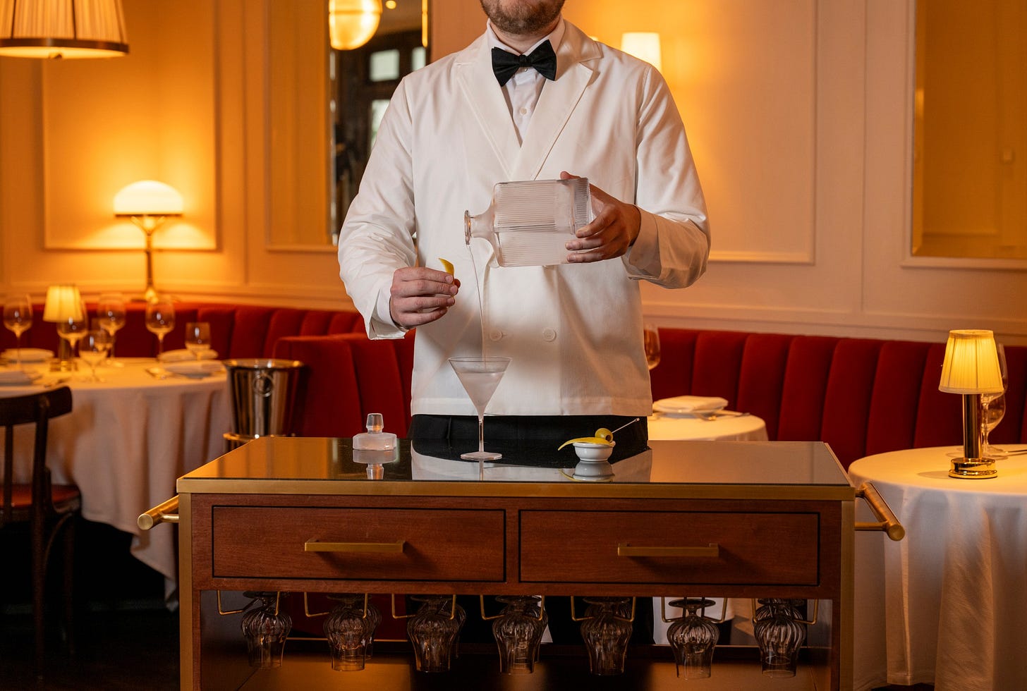 A bartender in a white tuxedo pouring a cocktail into a glass on a cart.