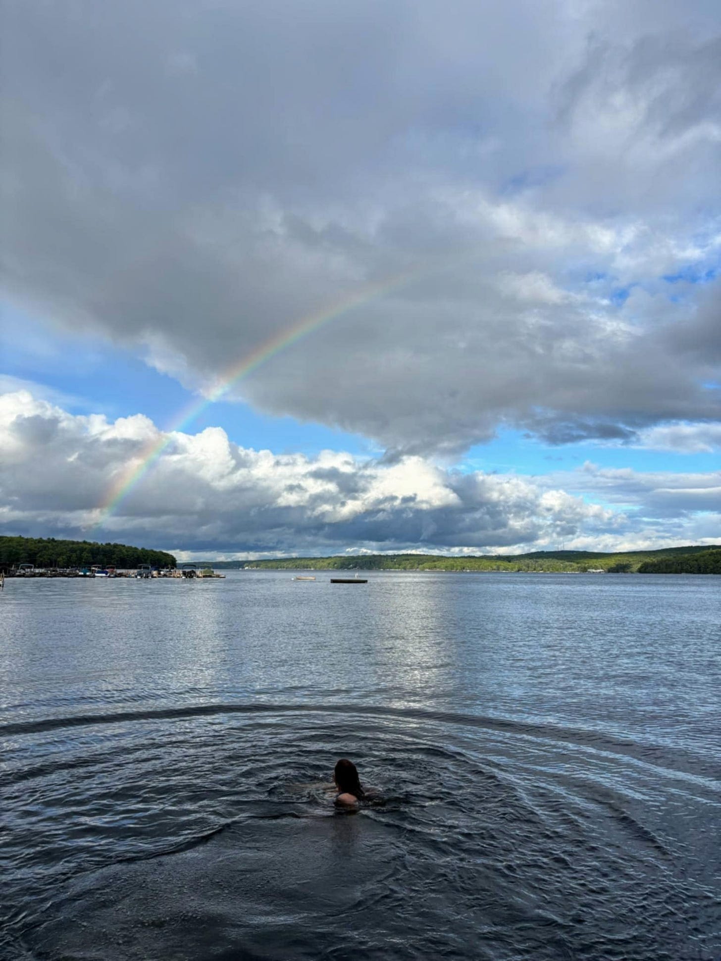 A person is swimming in a lake under a cloudy sky with a faint rainbow in the background. Trees line the shore, and docks with boats are visible on the left.