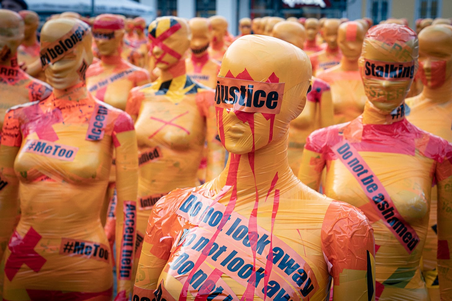 Imagem de protesto contra a violência de gênero. Manequins femininos envelopados com plástico amarelo, enrolados com fita plástica laranja com palavras-chave como #MeToo, stalking e justice, com adesivos vermelhos em forma de xis. Imagem de protesto contra a violência de gênero. Manequins femininos envelopados com plástico amarelo, enrolados com fita plástica laranja com palavras-chave como #MeToo, stalking e justice, com adesivos vermelhos em forma de xis.