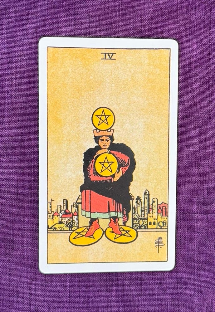 Tarot card on a purple linen cloth depicts a dark haired man sitting on a stool, beyond the boundaries of his hometown. His arms are wrapped tightly around a coin as if he fears he may lose it if he loosens his grip. He balances another pentacle on his head, and two more sit securely underneath his feet. He clearly doesn’t want anyone to take his coins. 