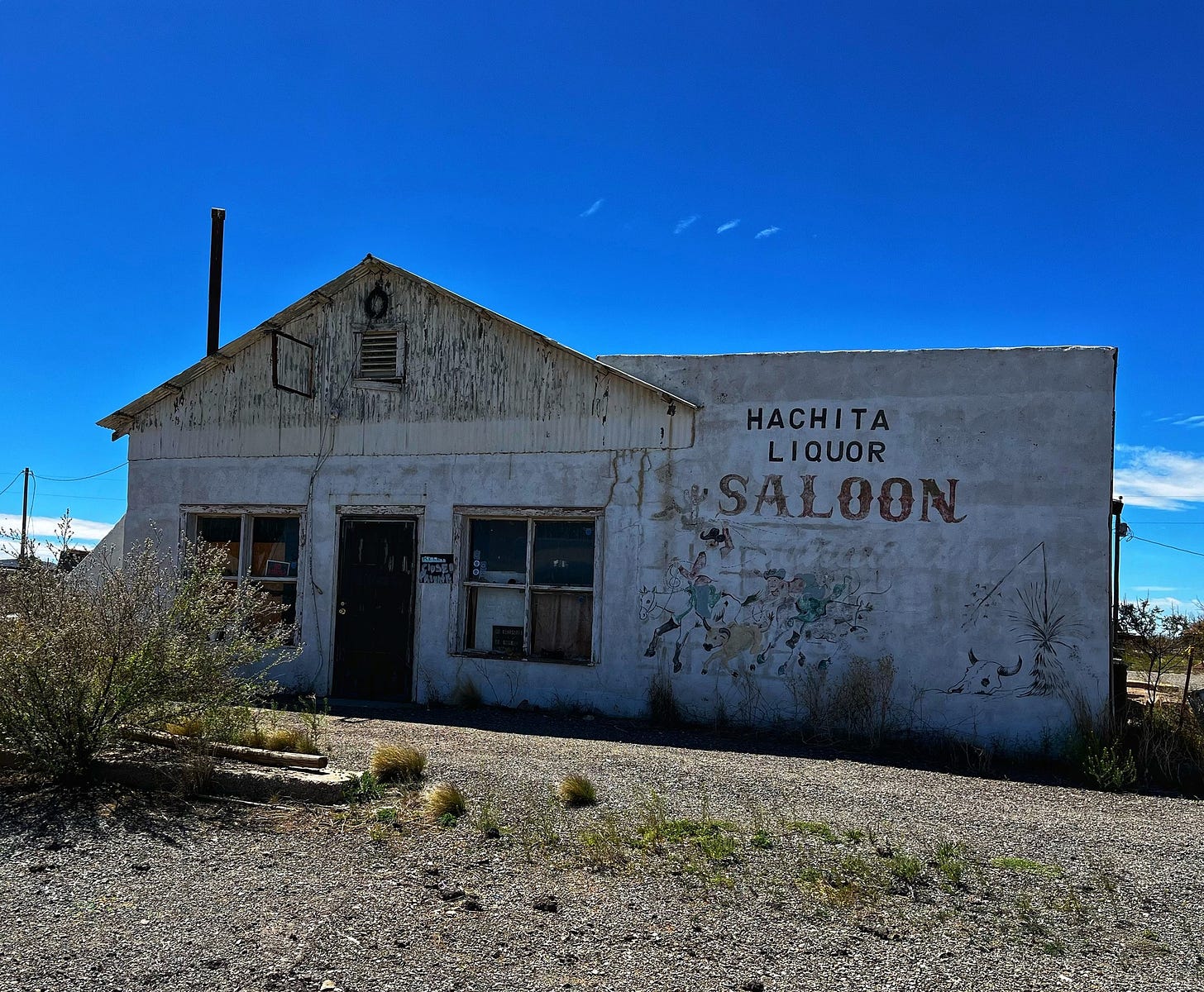 An abandoned saloon in a white building with blue sky behind. Painted letters on the building read Hachita Liquor Saloon.