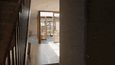 First-person view walking from a dim hallway past a wooden staircase into a bright open-plan living area with light wood floors, a small side table and chairs, and large glass doors opening to an outdoor space. First-person view walking from a dim hallway past a wooden staircase into a bright open-plan living area with light wood floors, a small side table and chairs, and large glass doors opening to an outdoor space.