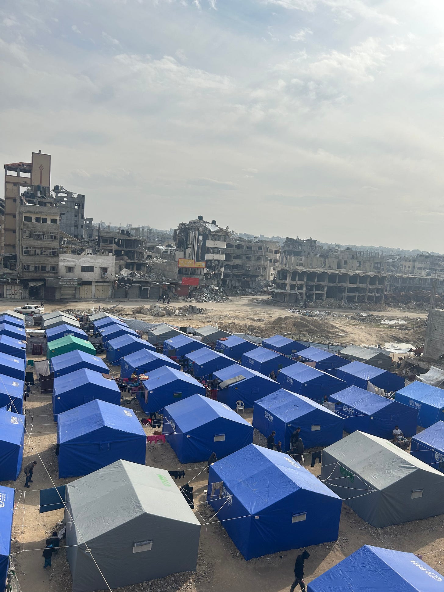 A wide view of a heavily damaged urban area with multiple large concrete buildings collapsed into piles of rubble under a gray, overcast sky. In the foreground, green shrubs and broken concrete frame the scene. A dirt road winds through the destruction, where a white van, a few cars, and several pedestrians are visible, highlighting ongoing activity despite the devastation. Makeshift shelters and scattered debris line the roadside, while a partially standing multi-story building rises in the background amid the ruins. A wide view of a heavily damaged urban area with multiple large concrete buildings collapsed into piles of rubble under a gray, overcast sky. In the foreground, green shrubs and broken concrete frame the scene. A dirt road winds through the destruction, where a white van, a few cars, and several pedestrians are visible, highlighting ongoing activity despite the devastation. Makeshift shelters and scattered debris line the roadside, while a partially standing multi-story building rises in the background amid the ruins.
