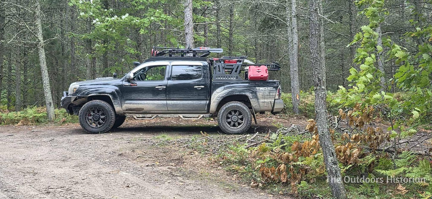 A black, second generation Toyota Tacoma with dirt on it from driving trails. It has a roof rack and racks over the box, with an extra gas tank, shovels, and other gear attached. It is parked in a dirt parking space off a dirt road with a forest behind it.